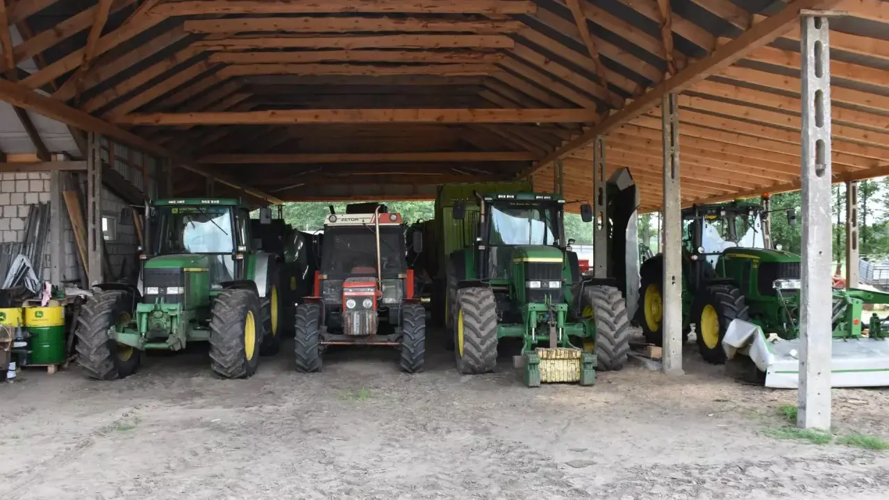 Kilka zielonych i jeden czerwony traktor stoją pod drewnianą wiatą. To pewnie ich tymczasowe schronienie, bo taka wiata bez pozwolenia to nie najlepszy pomysł.