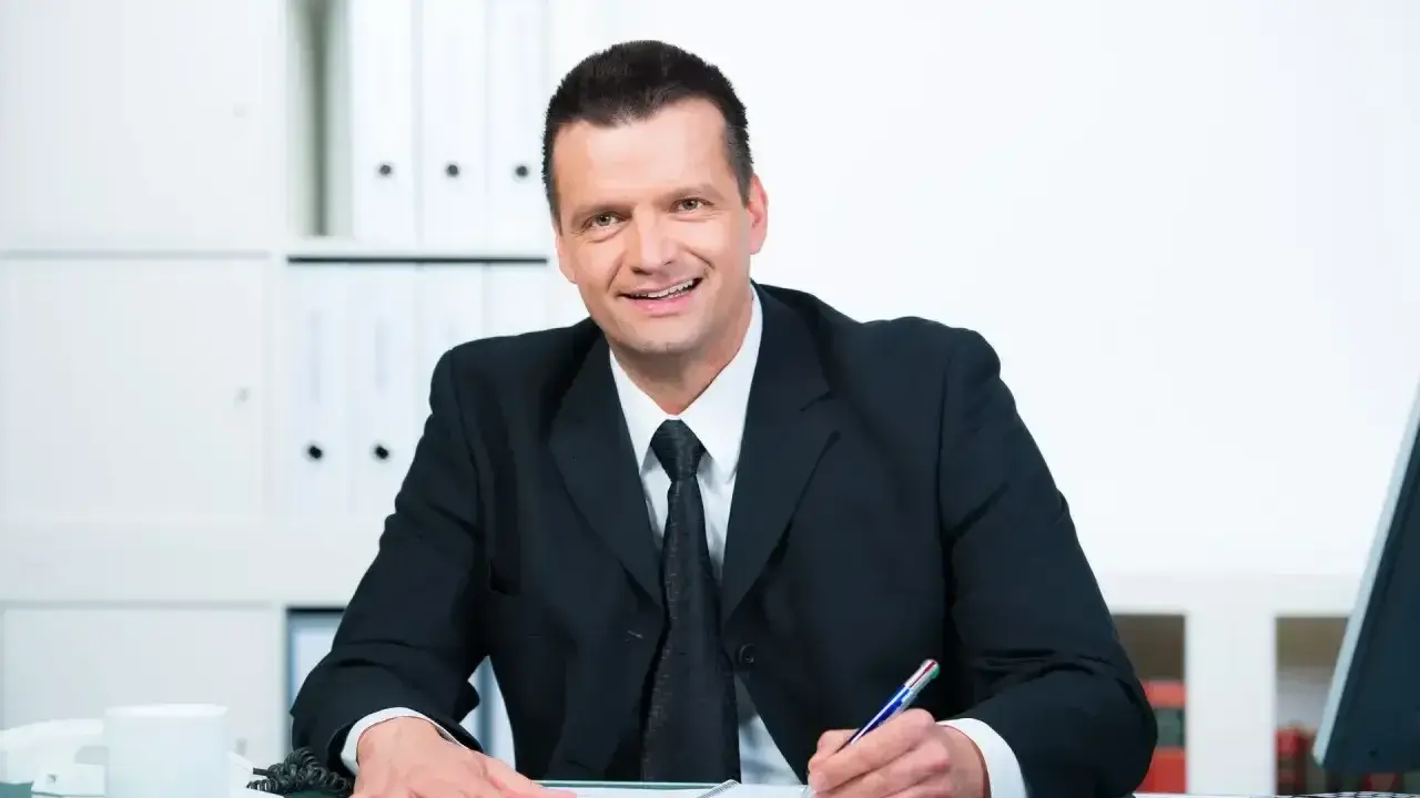 Uśmiechnięty mężczyzna w garniturze pisze w notatniku, przygotowując się do konkursu na dyrektora szkoły.