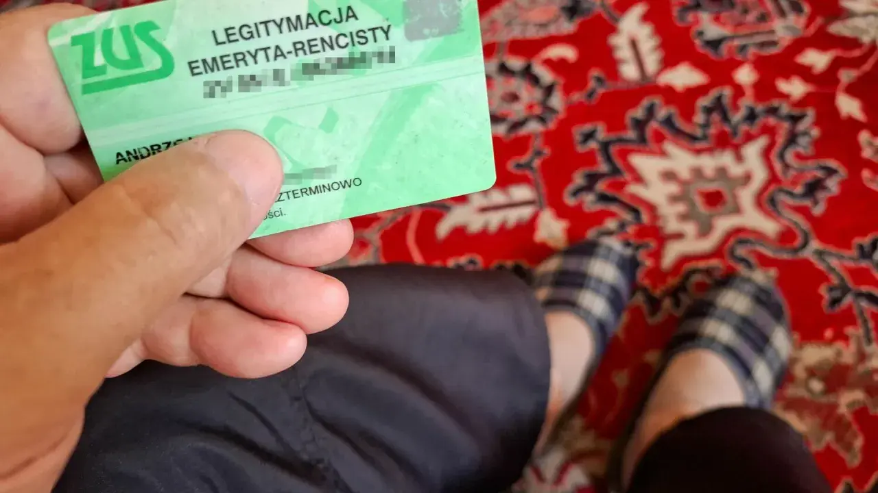 Zielona legitymacja ZUS "LEGITYMACJA EMERYTA-RENCISTY" w dłoni. Na dywanie widać stopy w kapciach.