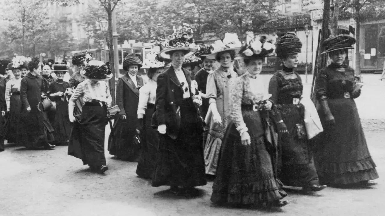 Frauen in langen Kleidern und Hüten marschieren entschlossen. Ein historisches Bild der feministischen Bewegung.