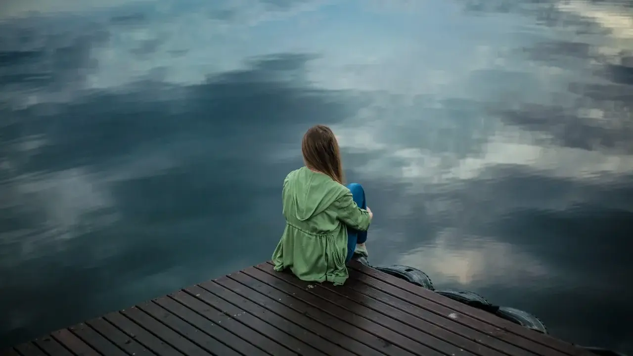 Eine Frau sitzt allein auf einem Steg, blickt aufs Wasser. Die Spiegelung der Wolken im Wasser verstärkt das Gefühl der **Angst vor dem Alleinsein**.