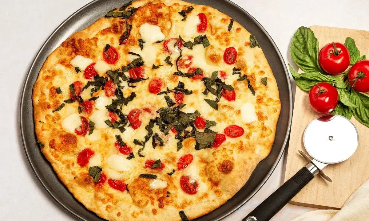 Pyszna pizza bez drożdży z pomidorkami koktajlowymi i mozzarellą, ozdobiona świeżą bazylią. Obok leży nóż do pizzy i składniki.
