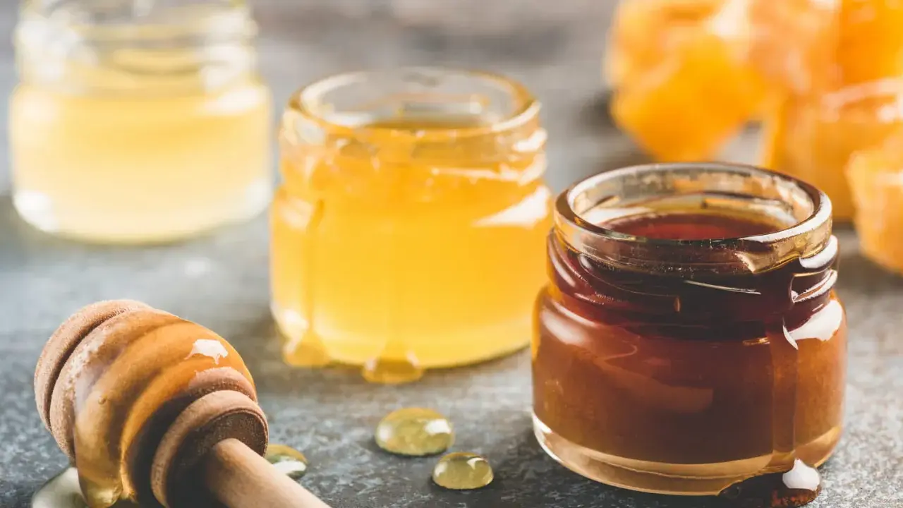 Trois pots de miel aux teintes variées, du clair au foncé, et une cuillère en bois dégoulinante. Quel miel pour pain d'épices ? Le choix est là !