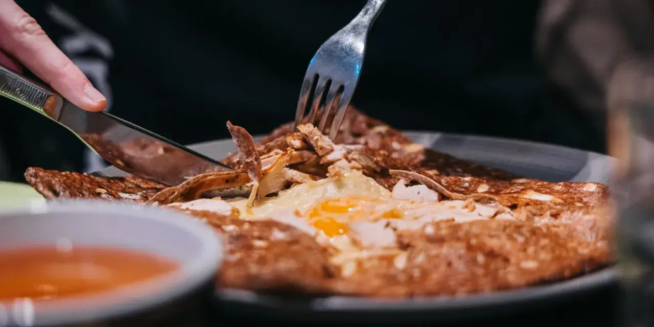 Une galette garnie d'œuf et de poulet, un délice des spécialités de Bretagne, est coupée avec un couteau et une fourchette.