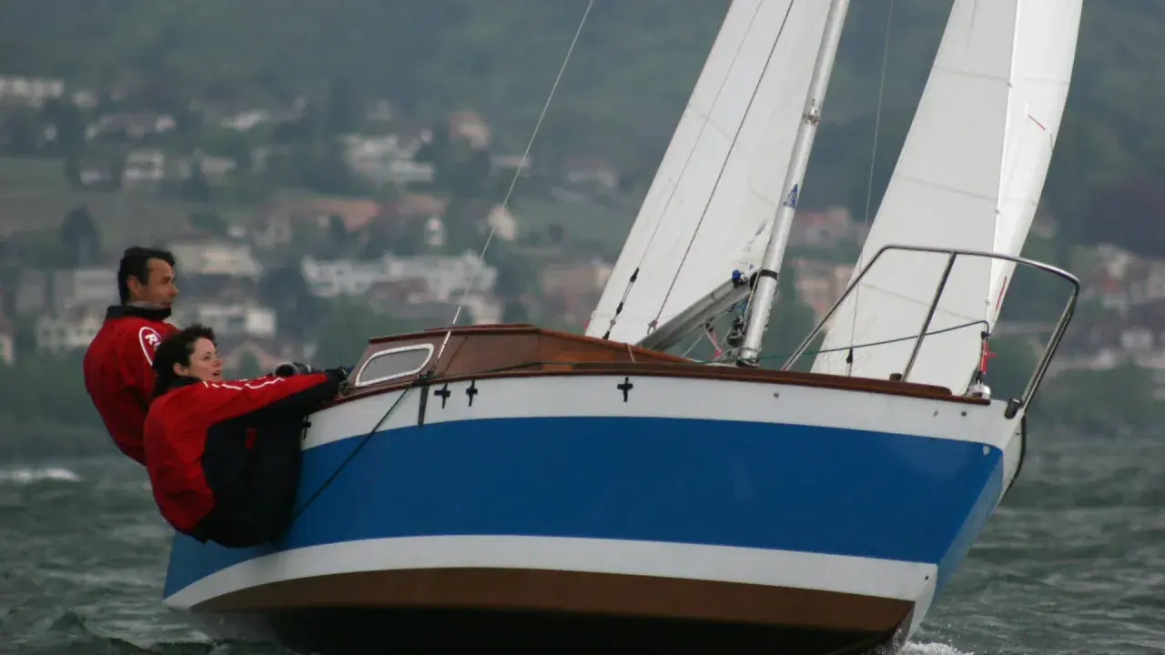 Couple en action sur un voilier transportable, naviguant sur des eaux agitées avec un village en arrière-plan.