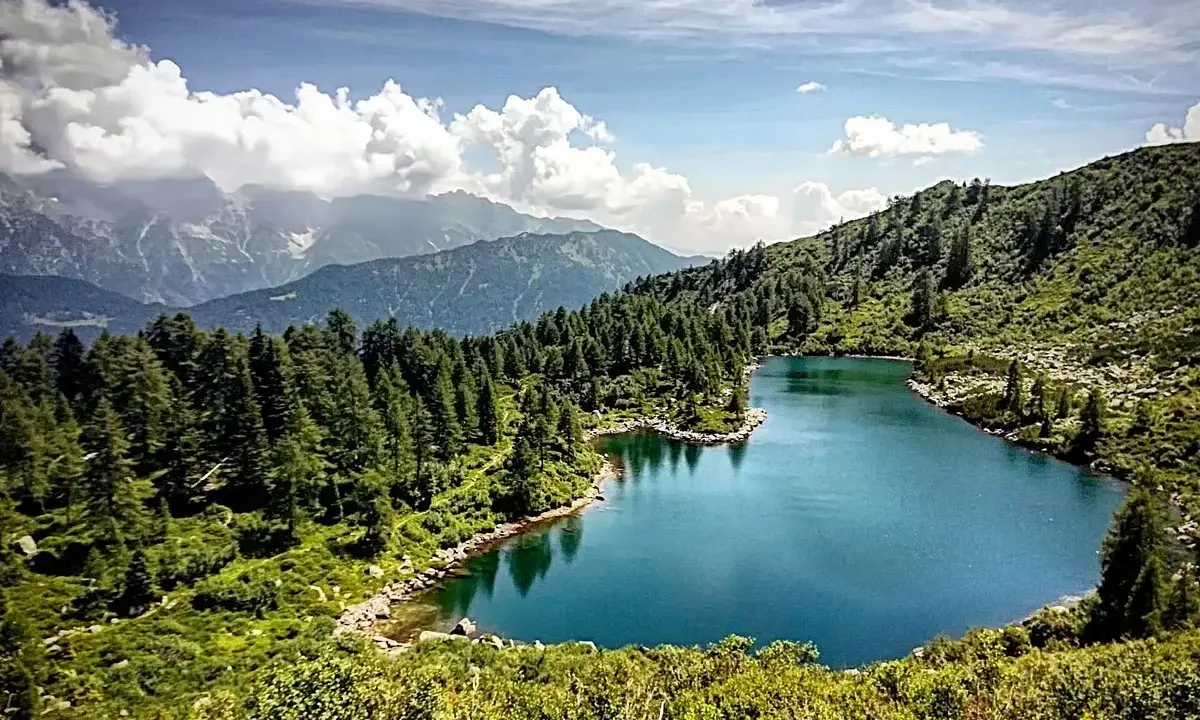 Laghi di San Giuliano: un'oasi alpina con acque turchesi, circondata da fitti boschi di conifere e montagne maestose sotto un cielo azzurro con nuvole bianche.
