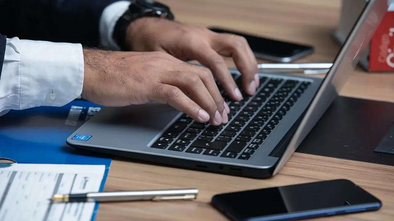 Prawnik pisze na laptopie, szukając informacji, jak napisać apelację od wyroku karnego wzór. Na biurku leżą dokumenty i telefon.