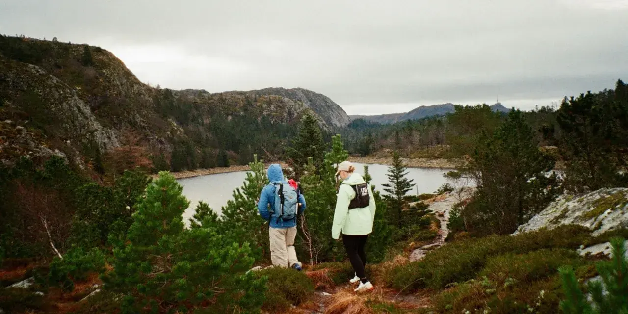 Två personer vandrar längs en stig med en sjö och berg i bakgrunden. Perfekt för att upptäcka Sveriges vandringsleder.