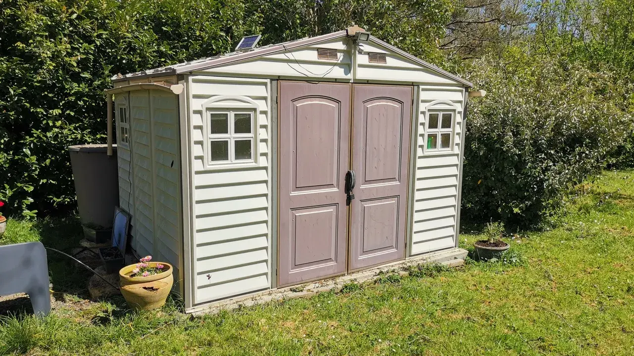 Un abri de jardin en plastique beige, avec des fenêtres et une double porte, est prêt pour son aménagement. Un petit panneau solaire est installé sur le toit.