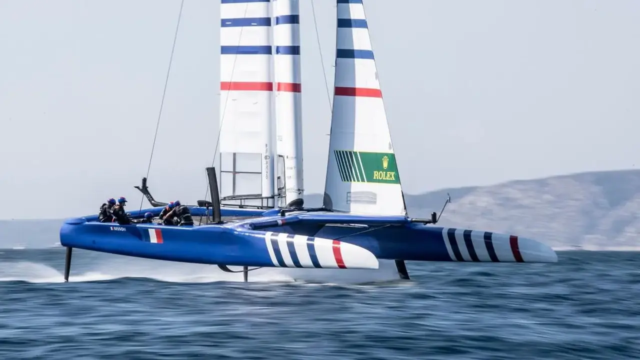 Catamaran bleu et blanc, voiles rayées, en pleine action. La **vitesse catamaran** est impressionnante, les foils hors de l'eau.