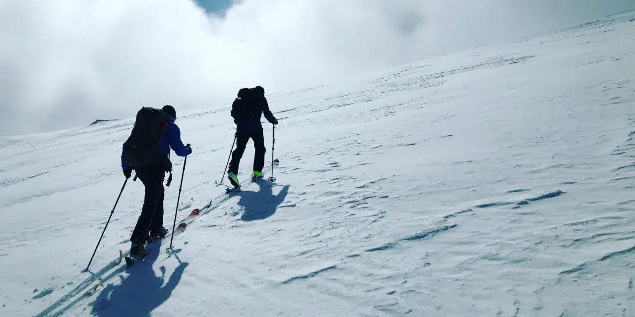 Två personer vandra på vintern uppför en snöklädd sluttning med skidutrustning och ryggsäckar.