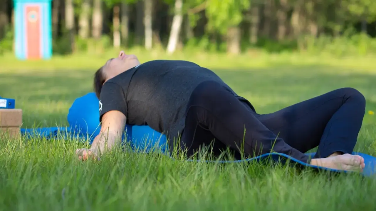 Person ligger på en blå yogarulle i gräset, med böjda knän och armarna utåt. En lugn stund med yin yoga utomhus.
