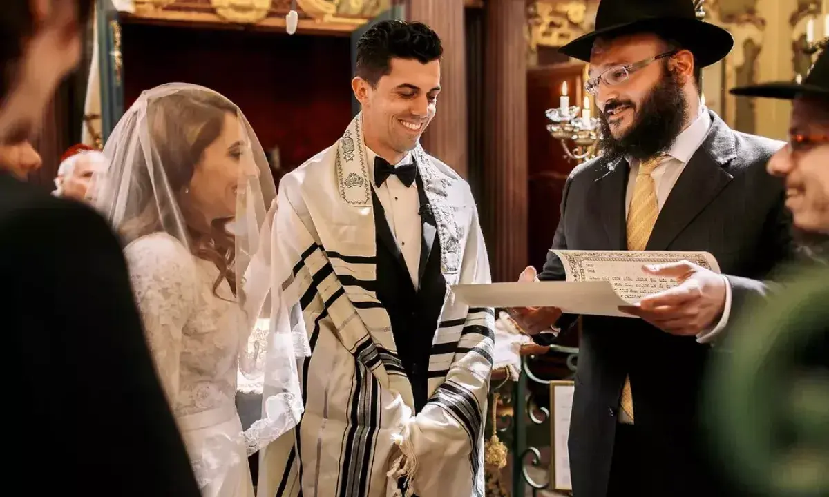 Novios sonríen durante su **matrimonio judío**. El novio lleva un talit y el rabino lee un documento.