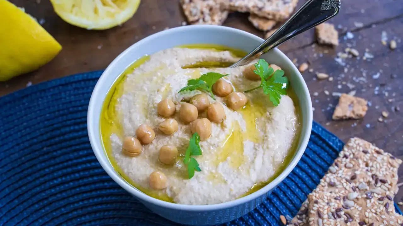 Cremiger Hummus, garniert mit Kichererbsen und Petersilie, daneben Zitronenspalten und knusprige Cracker. Das ist das beste Hummus Rezept!