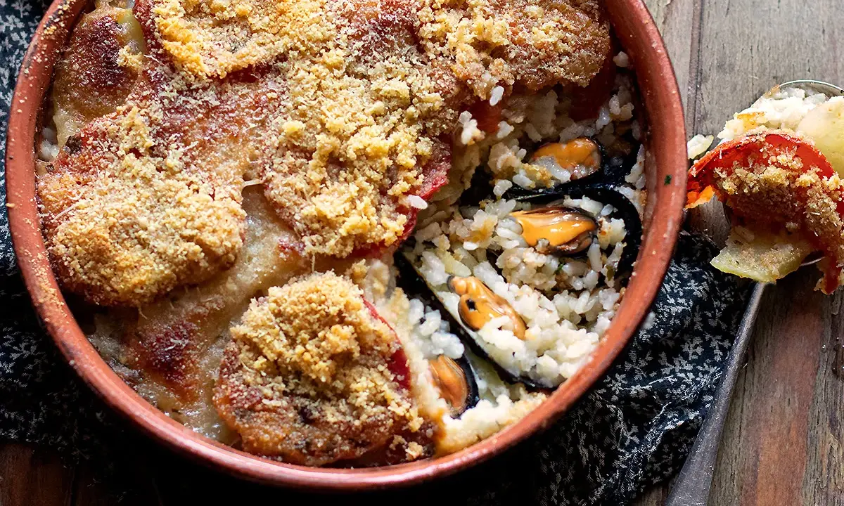 Piatto invitante di riso patate e cozze, gratinato con pomodoro e pangrattato, servito in una terrina di terracotta.