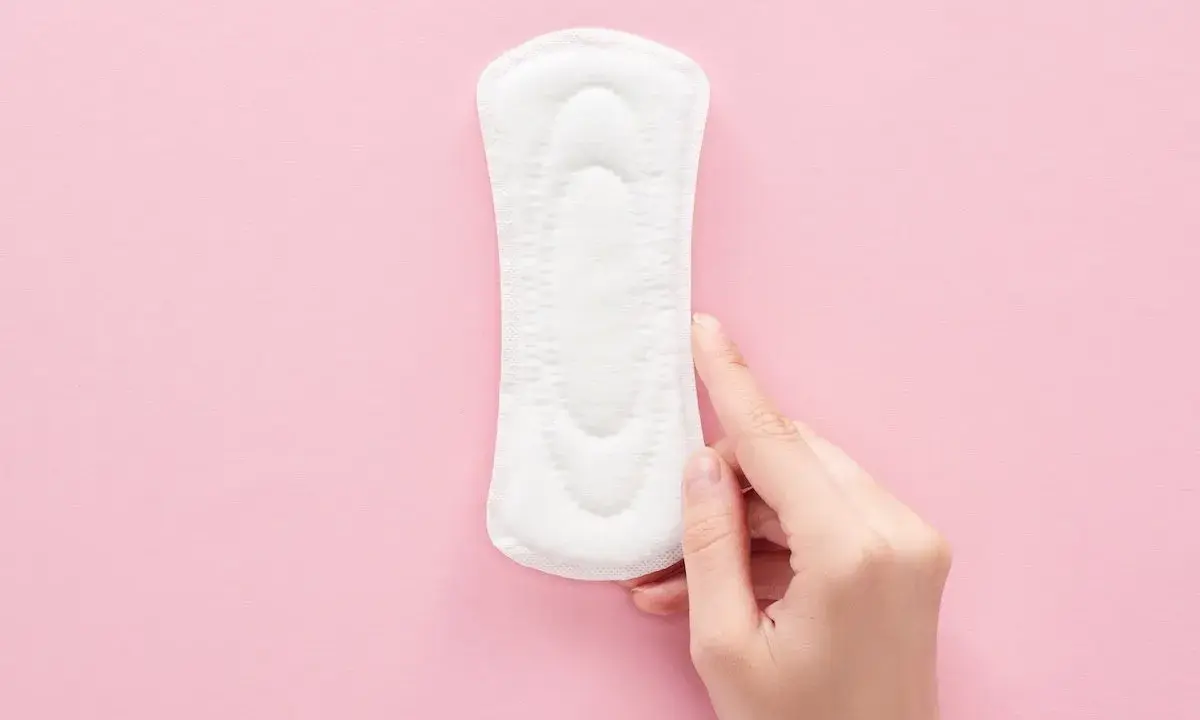 Una mano toca una compresa sobre fondo rosa. Representa cómo es el flujo antes de la regla, ligero y discreto.