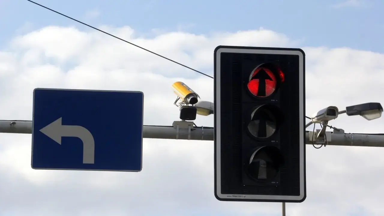 Rejestrator przejazdu na czerwonym świetle, widoczny na tle nieba, wraz z sygnalizacją świetlną i znakiem nakazu skrętu w lewo.