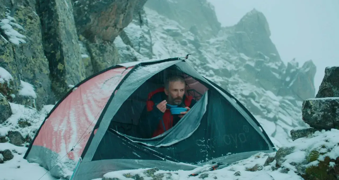 Mężczyzna w namiocie, jedzący posiłek, w zimowej scenerii górskiej. Scena jak z filmów wspinaczkowych.