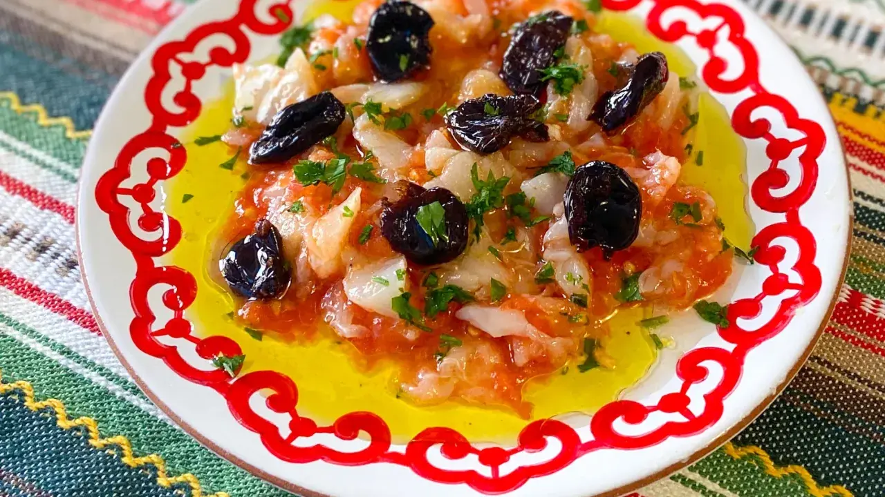 Esqueixada de bacalao con aceitunas negras y perejil, servida en un plato blanco con borde rojo sobre un mantel colorido.