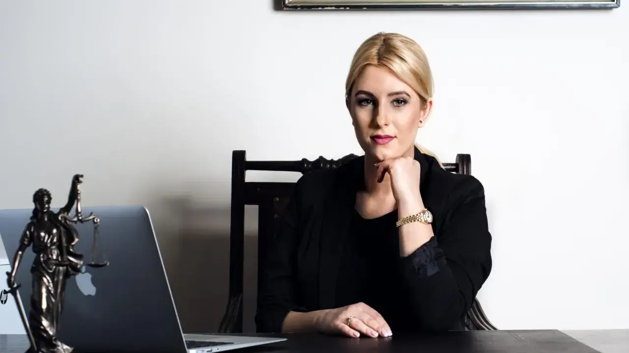 Kobieta w czarnej marynarce siedzi przy biurku z laptopem i figurką Temidy.