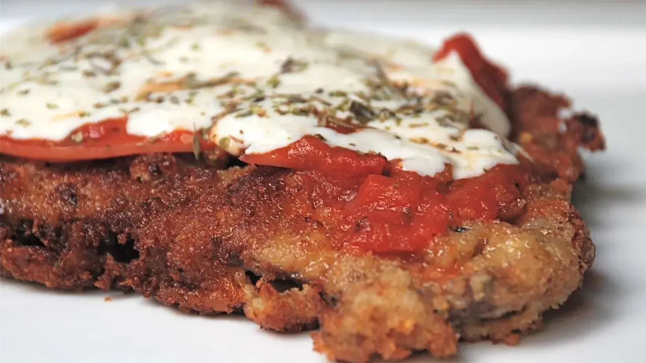 En närbild på en gyllene milanesa napolitana med smält ost, tomatsås och örter.