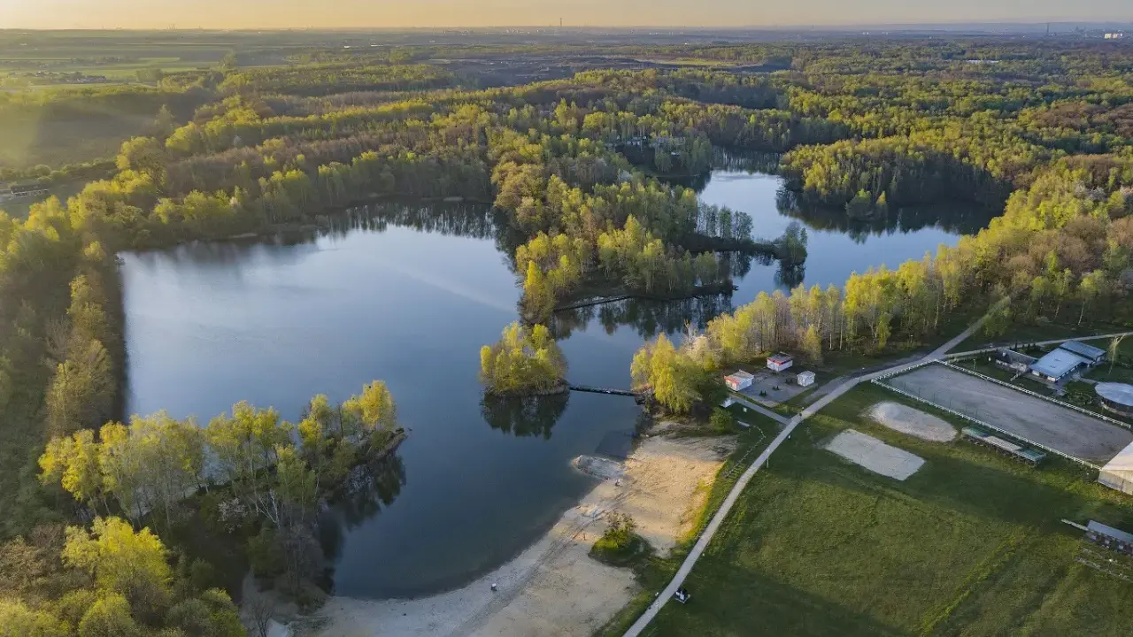 Malownicze jezioro Czechowice otoczone zielonym lasem, z piaszczystą plażą i zabudowaniami rekreacyjnymi.