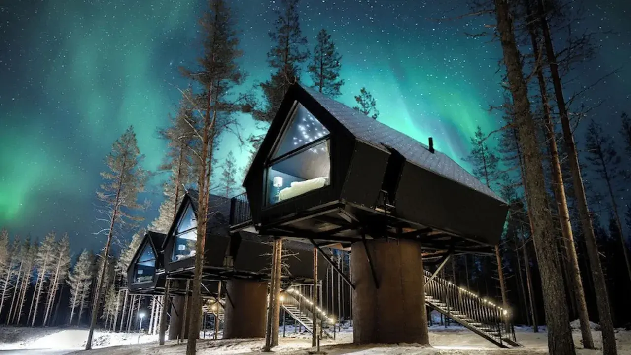 Hus på styltor i snöig skog under norrsken. En **arctic road** leder fram till stugorna, upplysta av stjärnor och lampor.