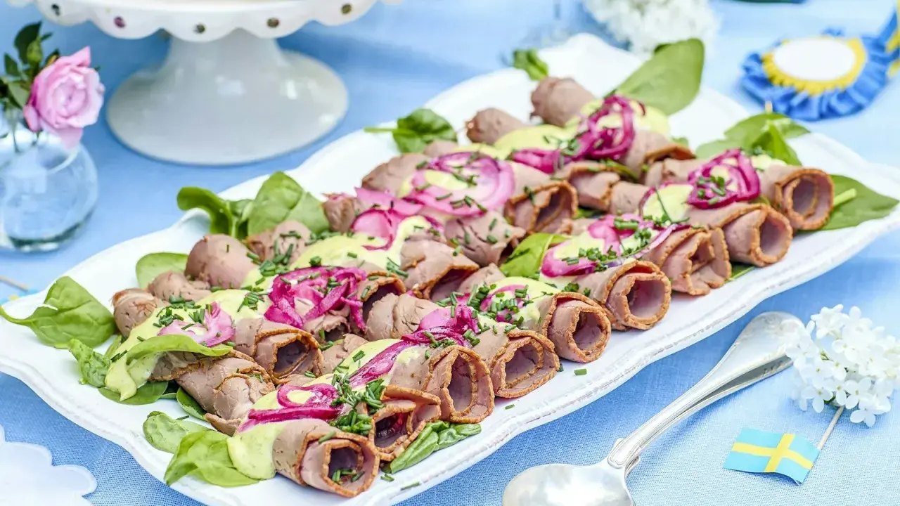 Rullad rostbiff med grön sås och rödlök, perfekt som enkel buffémat till många. Serveras på en vit bricka med gröna blad.