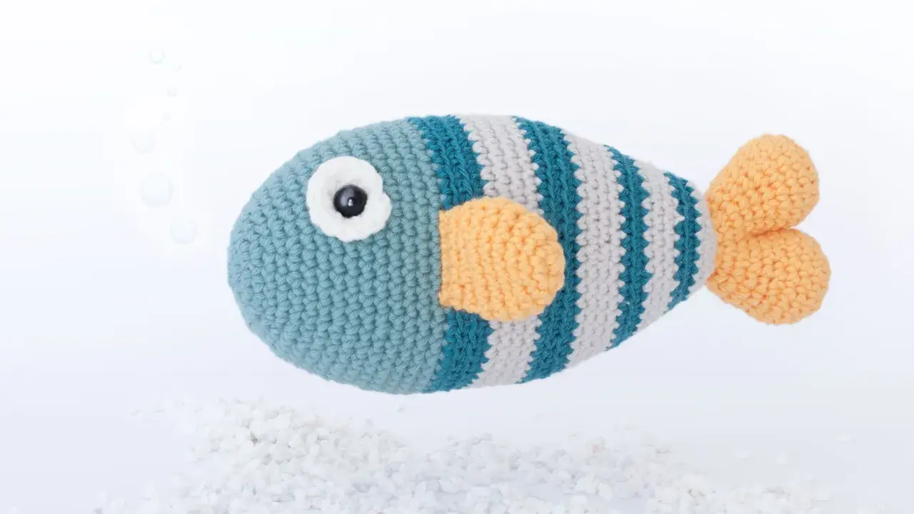 Un pez amigurumi a rayas azules y grises con aletas naranjas, flotando sobre guijarros blancos. ¡Encuentra aquí peces amigurumi patrón gratis!