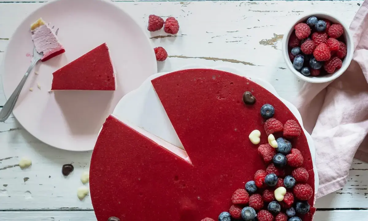 Ein köstlicher no bake Himbeer Cheesecake, dekoriert mit frischen Beeren und Schokoharz. Ein Stück ist bereits auf einem Teller.