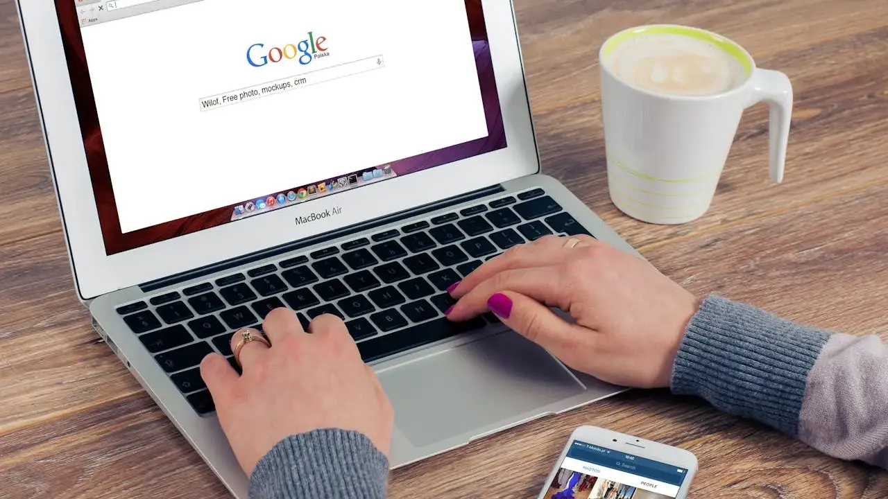 Kobieta pisze na laptopie z otwartą wyszukiwarką Google, obok stoi kubek kawy i smartfon.