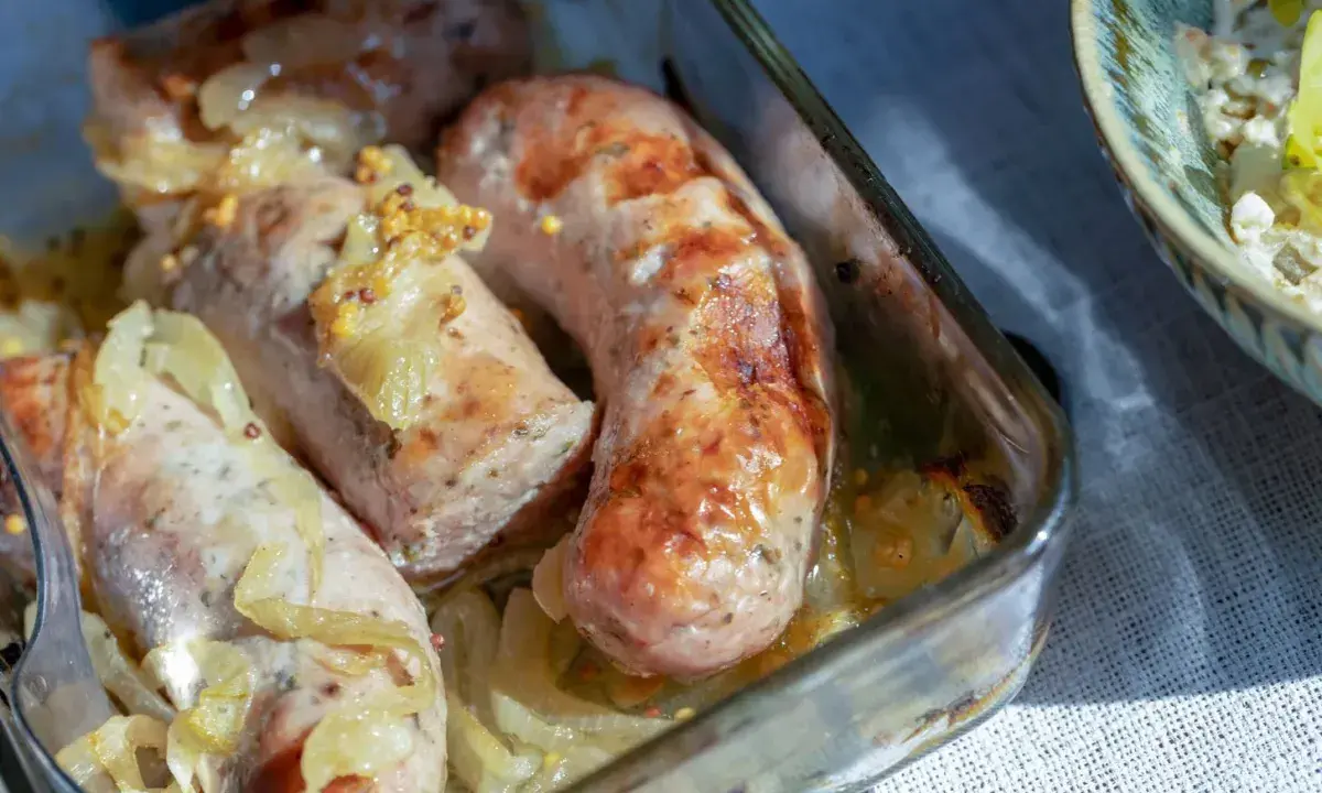 Soczysta biała kiełbasa pieczona z cebulką i musztardą, podana w szklanym naczyniu.