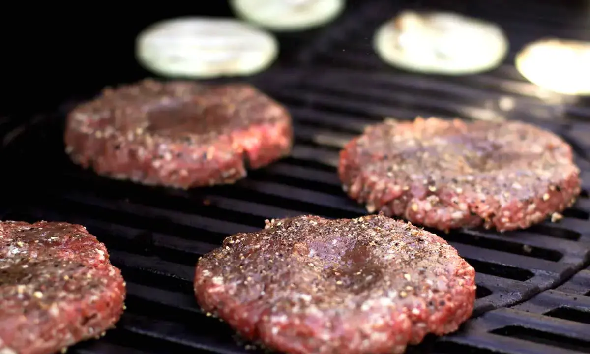 Vier frisch geformte Patties, gewürzt mit Pfeffer, brutzeln auf dem Grill. Perfekt, um Patties selber machen.