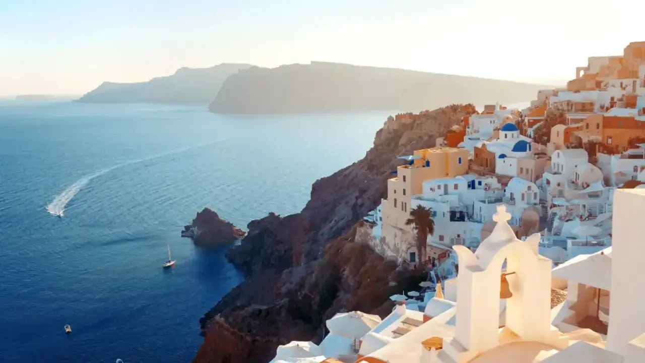 Traumhafter Blick auf die weißen Häuser und blauen Kuppeln von Santorini. Das Meer glitzert, Boote fahren vorbei. Ein perfekter Santorini Strand Urlaub.