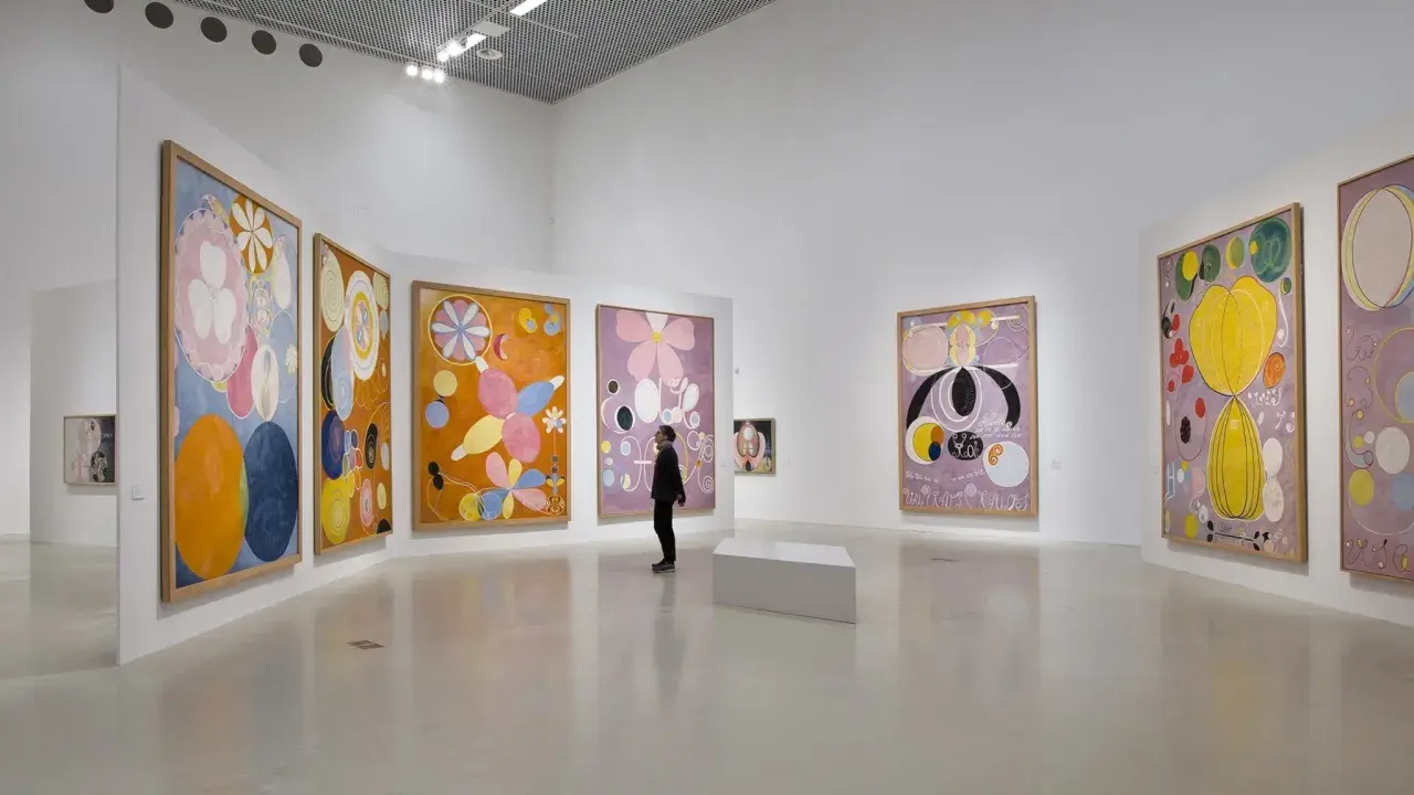 En person betraktar färgglada, abstrakta målningar av Hilma af Klint i ett ljust galleri.