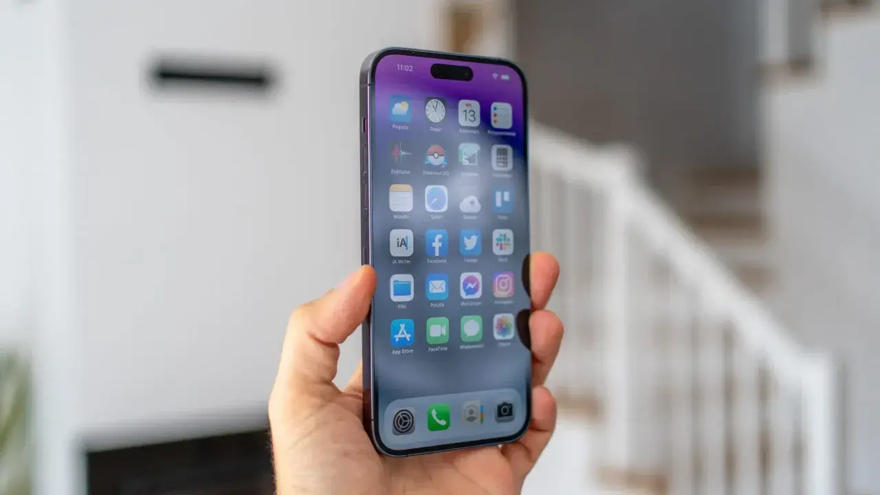 Dłoń trzyma smartfon z widocznym ekranem iPhone 14 Pro Max. Na wyświetlaczu widać ikony aplikacji i godzinę 11:02.