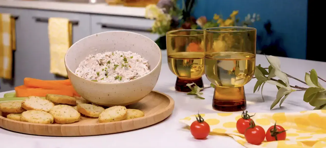 Un bol de rillettes de thon crémeuses, garni de ciboulette, servi avec des crackers, des bâtonnets de carottes et de céleri. Deux verres de vin blanc complètent cette scène gourmande.