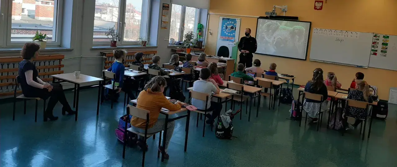 Dzieci słuchają lekcji o bezpieczeństwie w szkole. Nauczycielka i policjant prowadzą zajęcia.