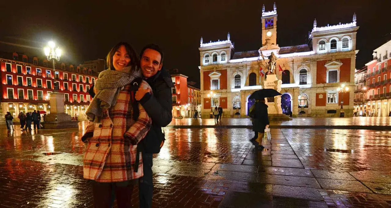 Pareja sonriente en la Plaza Mayor de Valladolid, disfrutando de las atracciones nocturnas y el reflejo de las luces en el suelo mojado.