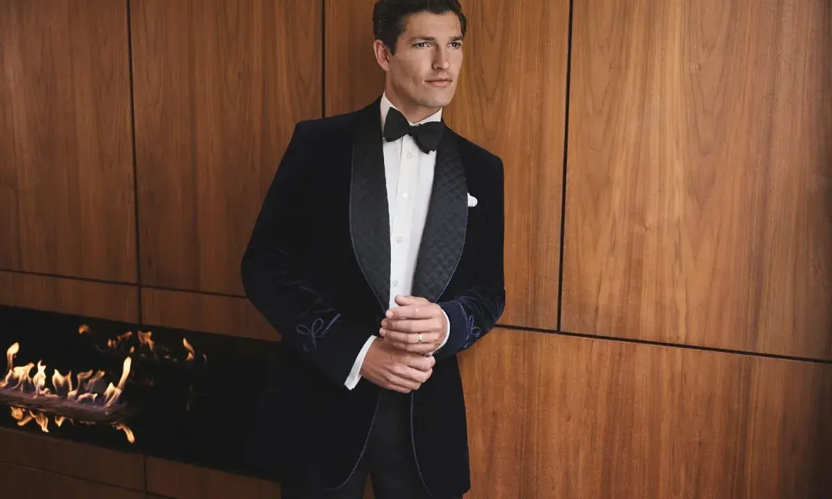 A man in a sophisticated navy velvet tuxedo, embodying perfect black tie attire, stands by a modern fireplace.