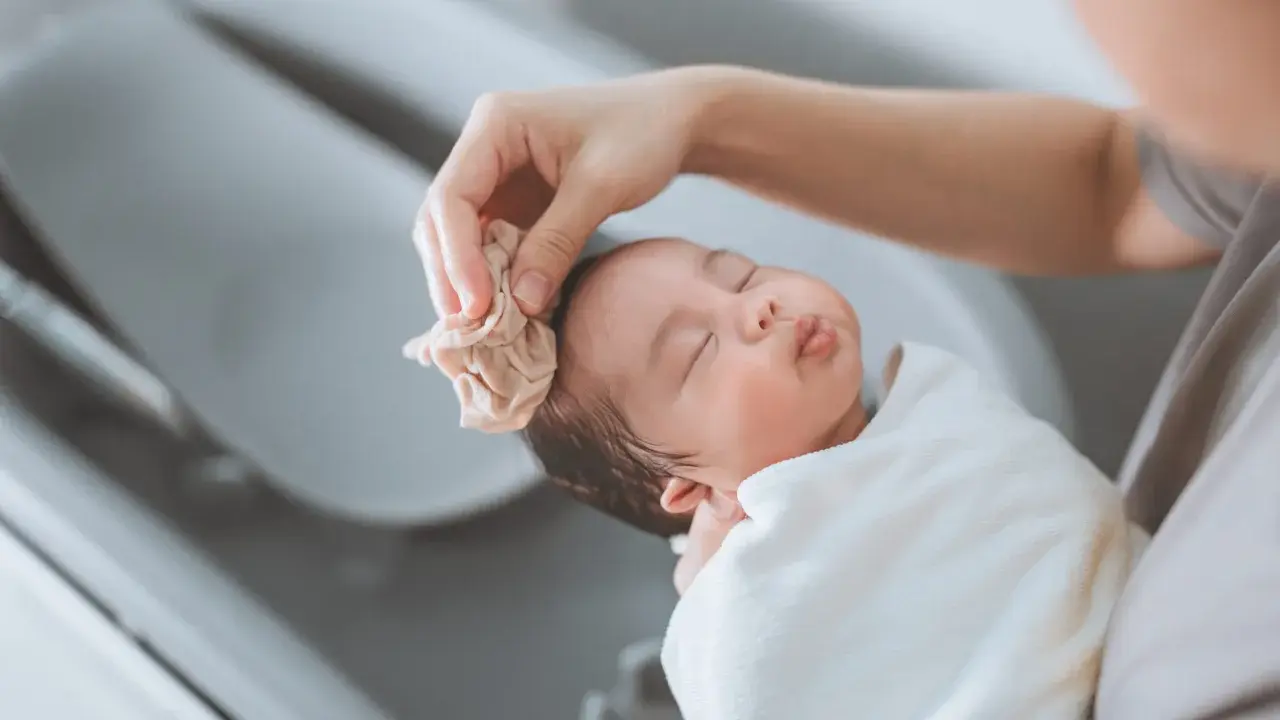 Bain d'un nouveau-né : une main nettoie délicatement la tête du bébé avec une petite serviette. Une scène paisible qui rappelle la liste produits toilette bébé naissance.