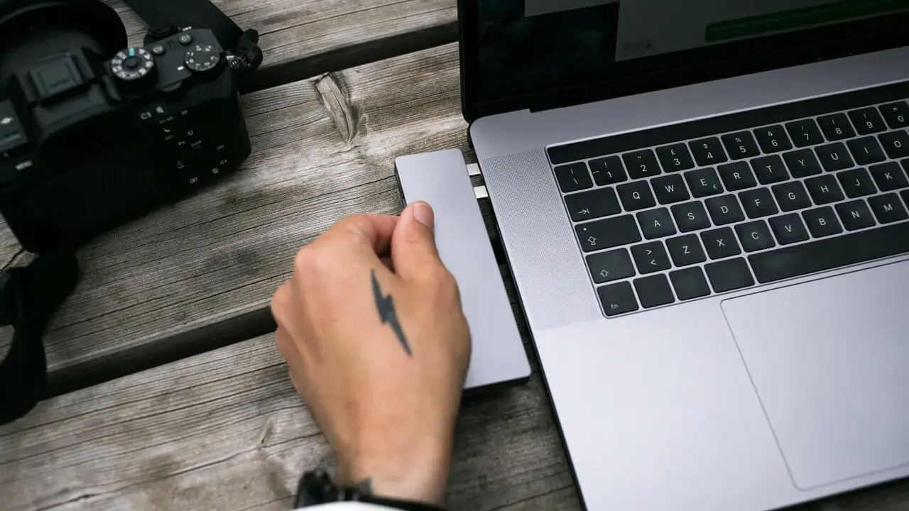 Dłoń z tatuażem pioruna podłącza adapter do laptopa, obok leży aparat. Idealny zestaw, jaki laptop do obróbki zdjęć?