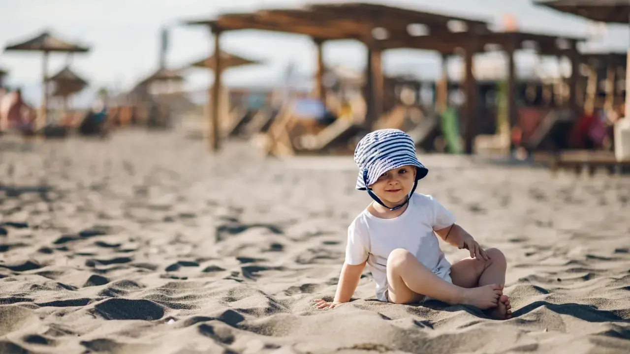 Uroczy maluch w pasiastym kapelusiku siedzi na piaszczystej plaży. Idealne miejsce, gdzie na wakacje z rocznym dzieckiem.