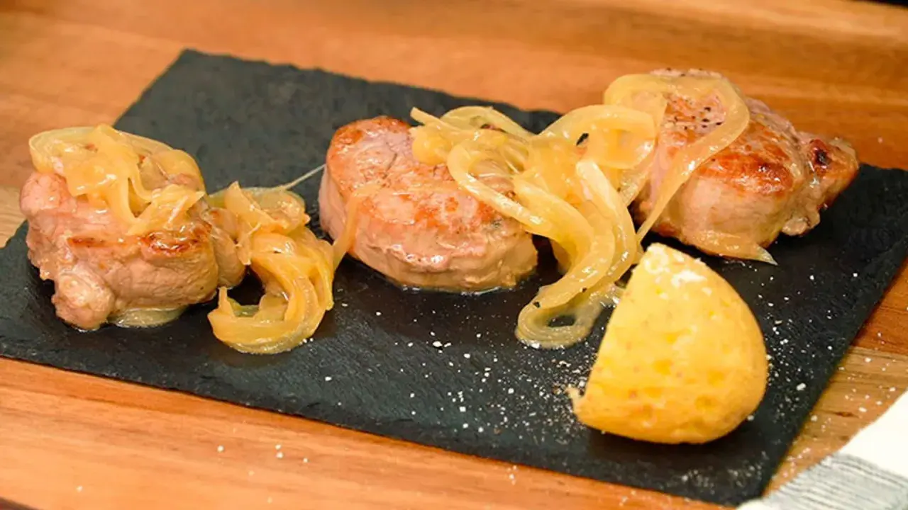 Tiernos medallones de carne al jerez, cubiertos con cebolla caramelizada y acompañados de una porción de patata.