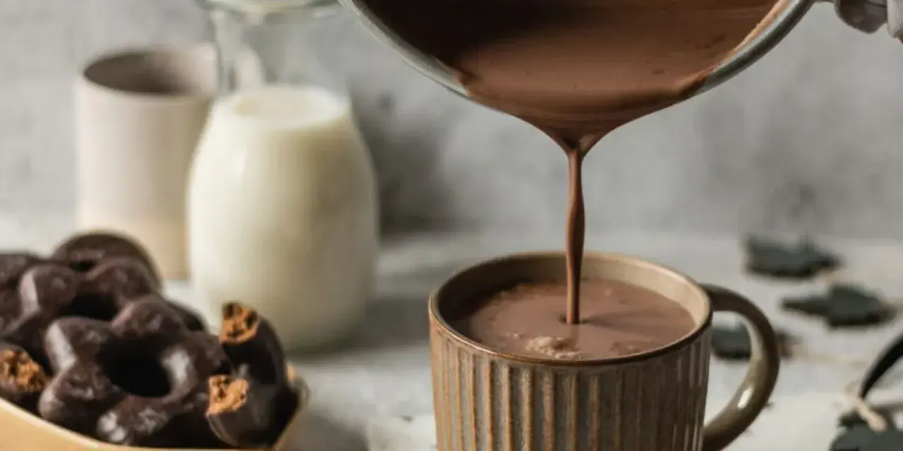 Varm choklad hälls upp i en mugg. Bredvid står en skål med chokladkakor och en mjölkflaska.