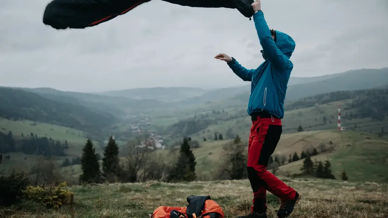 Osoba w niebieskiej kurtce i czerwonych spodniach rozkłada śpiwór puchowy czy syntetyczny na tle górskiego krajobrazu.