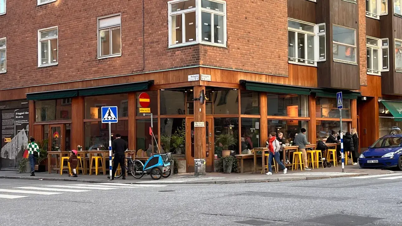 Ett mysigt hörnkafé i **Södermalm, Stockholm** med folk som sitter vid gula stolar och bord utomhus. En blå cykel med barnvagn står parkerad.