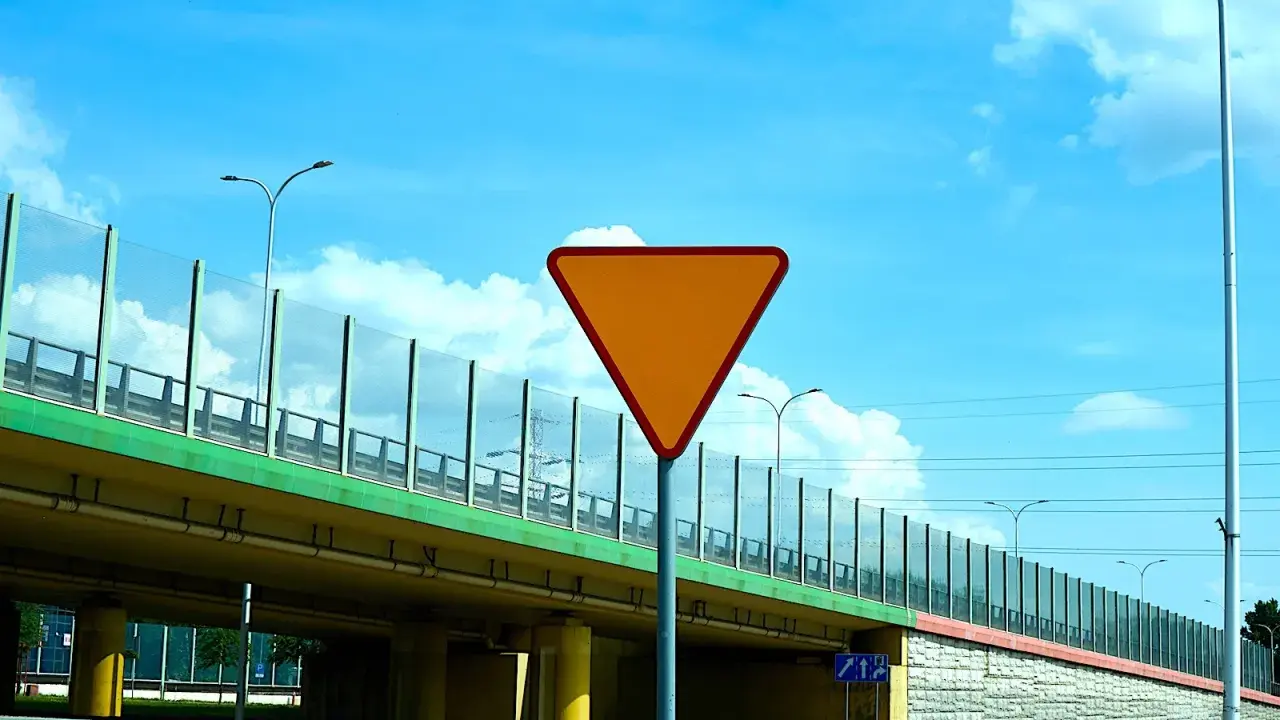 Pomarańczowy znak "ustąp pierwszeństwa" ostrzega przed wjazdem na drogę podporządkowaną. Pod wiaduktem widać drogę i słupy oświetleniowe.