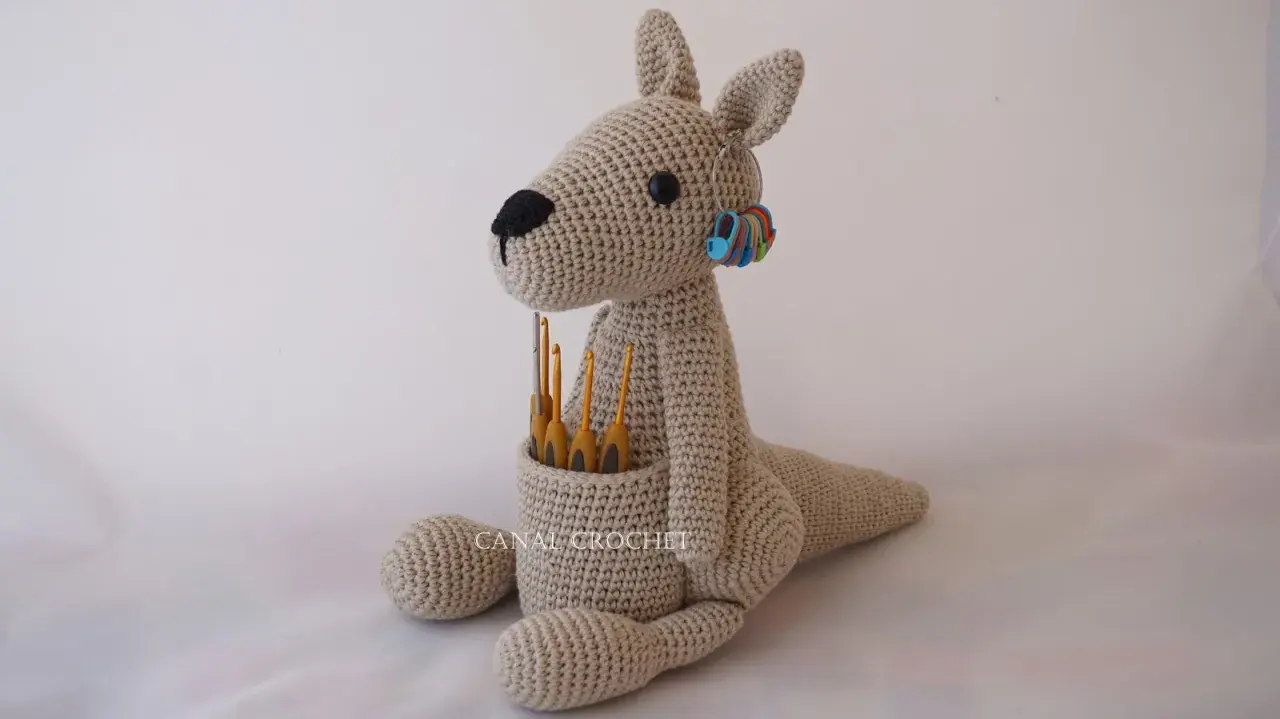 Canguro amigurumi beige sentado, con ganchillos en su bolsa y marcadores de puntos en su oreja.