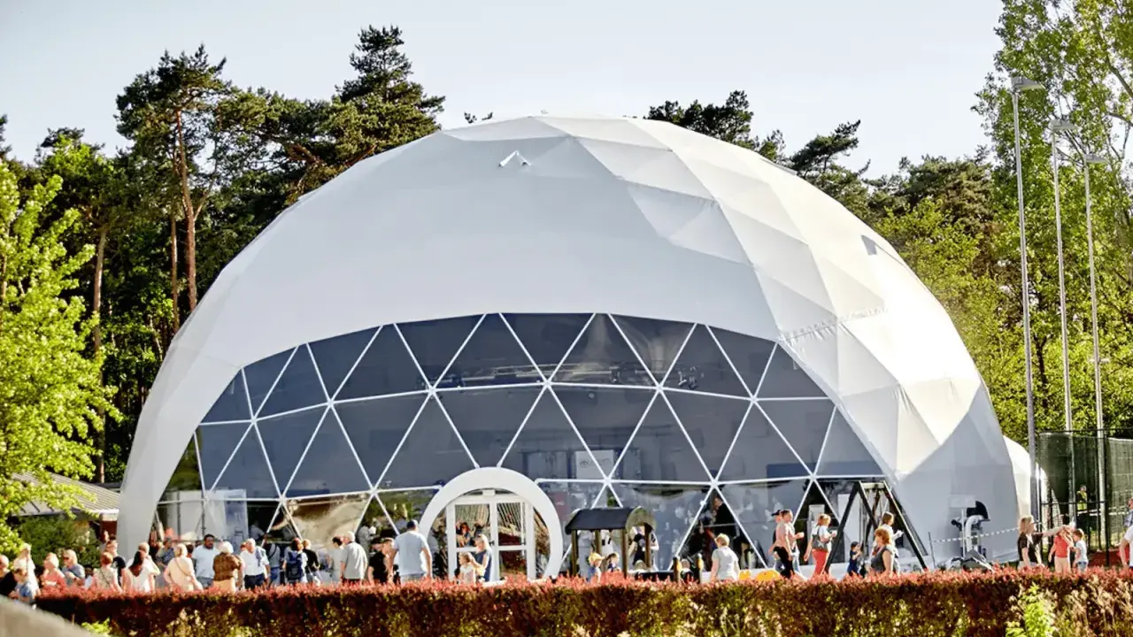 Una gran cúpula estructura blanca, con ventanas triangulares, se alza en un día soleado. Gente pasea frente a ella, rodeada de árboles verdes.